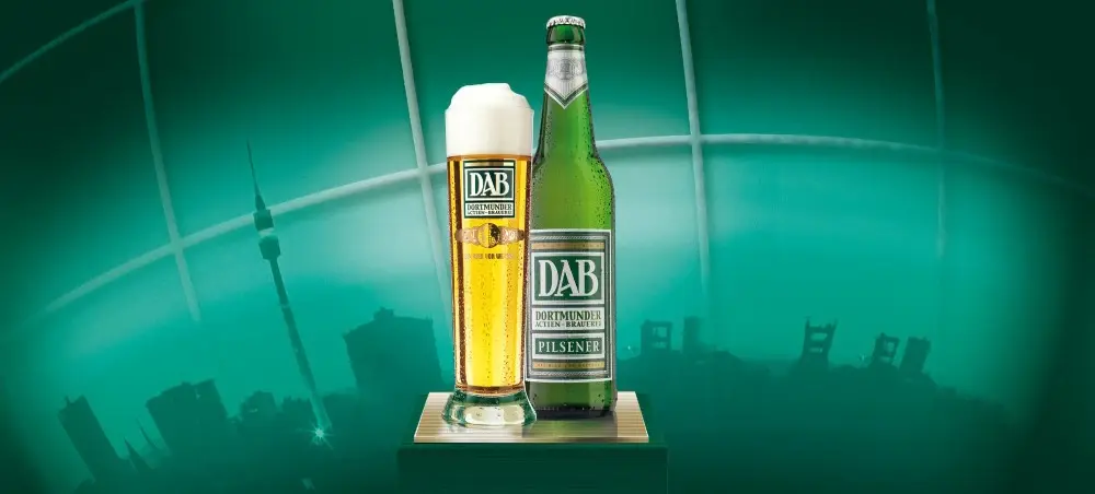 Eine gefüllte Bierglas steht neben einer grünen Bierflasche vor einer Stadtansicht.