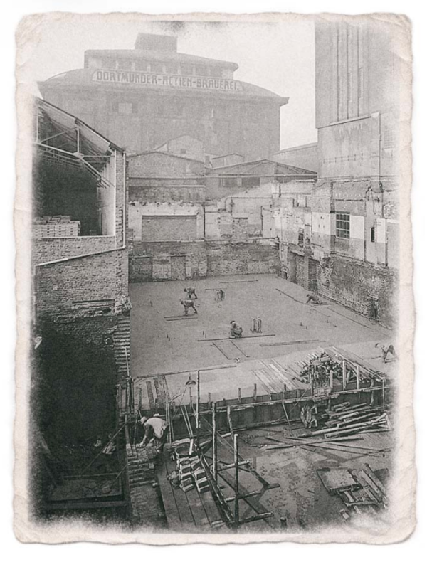 Historische schwarz-weiß Aufnahme einer Baustelle mit Arbeitern und Gebäuden im Hintergrund.