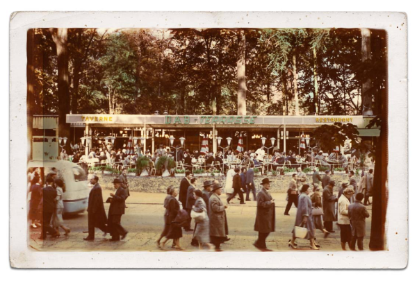 Leute gehen vor einem belebten Restaurant in einem Park spazieren, einige sitzen an Tischen.