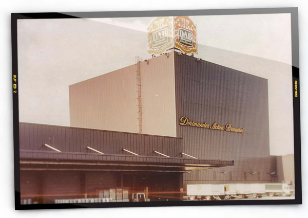 Brauereigebäude mit einer großen Fassade und Dominantem Logo. Auf der Ladefläche stehen LKW.